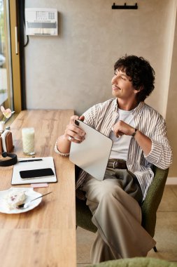 Genç bir adam bir kafede rahatça oturur, elinde bir not defteri tutarken huzur içinde yansıtır ve gülümser..