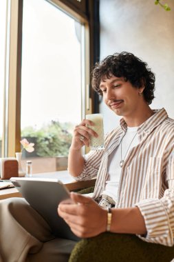 Yakışıklı genç adam bir kafede rahatça oturur, içkisini yudumlar ve gülümseyerek tablet kullanır..