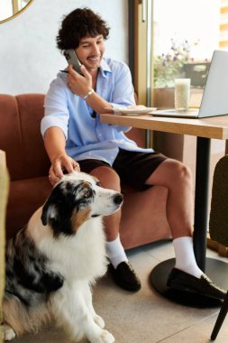 Moda bir kafede, yakışıklı genç bir adam telefonda konuşurken gülümsüyor, bir köpeği okşuyor..