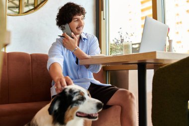Genç bir adam dizüstü bilgisayarı üzerinde çalışırken yanında bir köpekle telefon görüşmesi yapıyor..