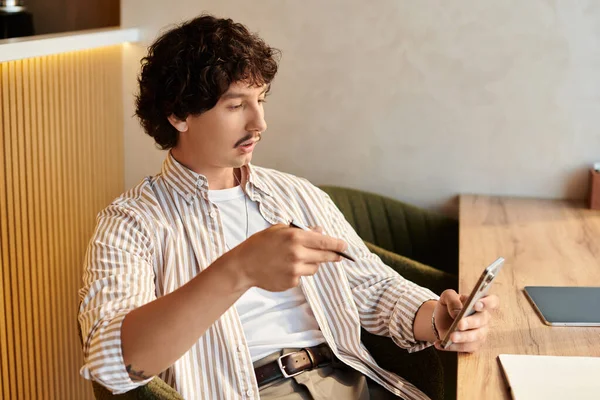 Genç bir adam şık bir kafede oturmuş elinde kalemle akıllı telefonuyla düşünceli bir şekilde çalışıyor..