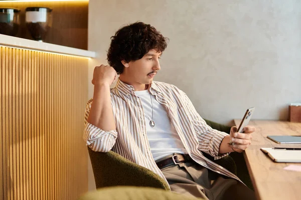 Yakışıklı genç bir adam modern bir kafede rahat bir şekilde oturuyor, akıllı telefonuyla meşgul..