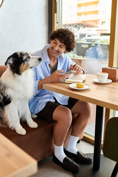 Young man smiles while savoring dessert and coffee, accompanied by his loyal dog at a cafe.