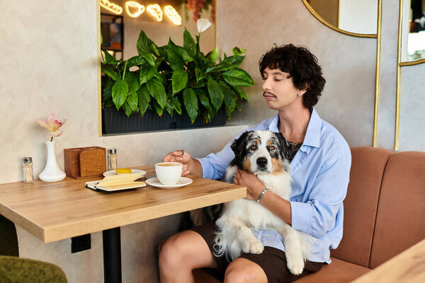 В уютном кафе красивый молодой человек расслабляется, потягивая кофе, лаская свою собаку с радостью.