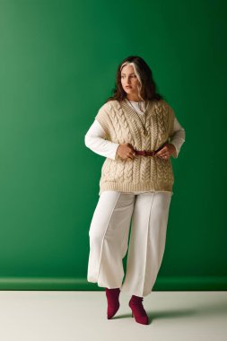 Rahat bir süveter ve şık bir pantolon giyen kendine güvenen bir kadın yeşil bir arkaplanda zarif bir şekilde duruyor..