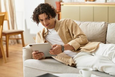 Şık giyimli adam kanepede uzanır, rahat bir yaşam alanında tabletle mutlu mesut meşgul olur..