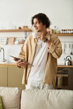 Modaya uygun genç bir adam kahvesini yudumlar ve modern bir mutfak ortamında tablet bulur..