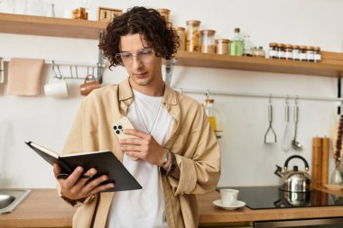 Yakışıklı genç adam şık bir mutfakta akıllı telefon tutarken defter okuyor..