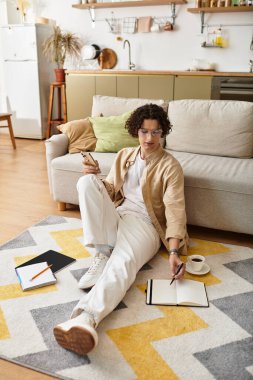 İş kıyafeti giymiş genç bir adam yere rahatça oturur ve fikirlerini yazar..