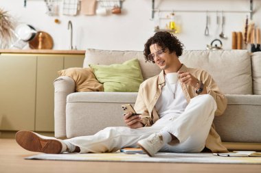 Yakışıklı genç adam yerde rahatça dinleniyor, telefonuyla meşgulken kahvesini yudumluyor..