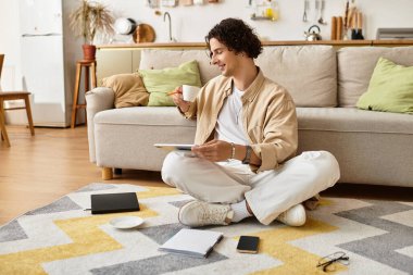 Yakışıklı genç adam bacak bacak üstüne atarak yerde oturuyor, kahvesini yudumluyor ve tabletine odaklanıyor..