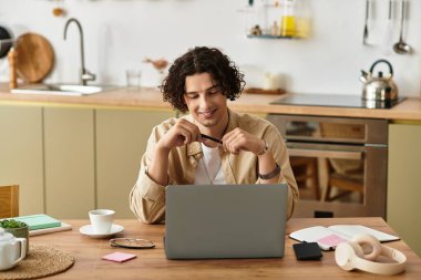 İş dünyasından yakışıklı genç bir adam modern bir mutfakta dizüstü bilgisayarıyla çalışırken gülümsüyor..