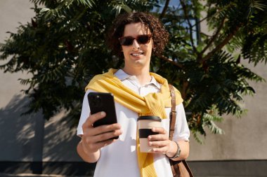 Şık giyinmiş yakışıklı genç adam elinde kahve fincanı ve gülümseyen bir akıllı telefon tutuyor..