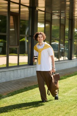 Şık giyinmiş genç bir adam bir çanta taşırken kahve fincanıyla mutlu bir şekilde yürür..