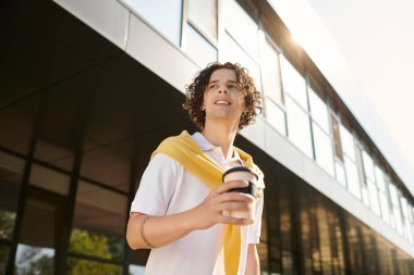 Şık giyinmiş yakışıklı genç adam elinde kahve tutarken gülümsüyor, güneşin tadını çıkarıyor..