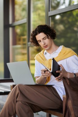 İş kıyafeti giyen yakışıklı genç adam dizüstü bilgisayar ve defterle özenle çalışır..