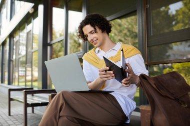 Çekici bir genç adam bilgisayarında çalışıyor ve modern bir açık havada otururken elinde bir tablet tutuyor..