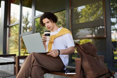 Takım elbiseli yakışıklı bir genç dışarıda laptopunu kullanırken kahvesini yudumluyor..