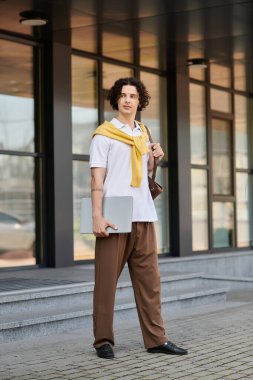 Modern iş kıyafeti giymiş yakışıklı genç adam ofis binasının yanında duruyor..