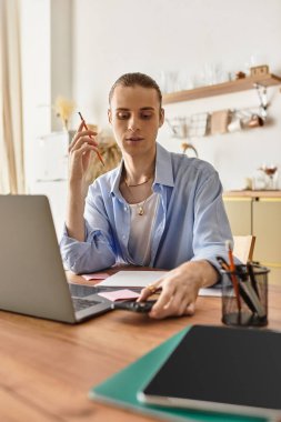Çekici, kadınsı bir genç adam masasında düşünceli bir şekilde çalışıyor. Etrafı renkli malzemelerle çevrili..