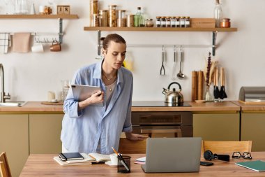 Genç yakışıklı bir adam şık bir mutfakta elinde tabletle dizüstü bilgisayarına odaklanmış duruyor..