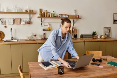 Genç bir kadın erkek, güneşli bir mutfak alanında, ahşap bir masada dizüstü bilgisayara odaklanıyor..