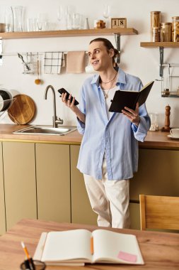 Samimi bir mutfakta, gülümseyen genç bir adam elinde bir telefon ve not defteri ile pozitiflik saçıyor..