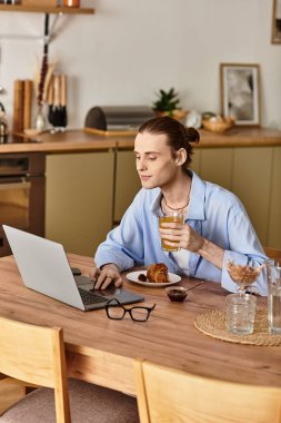 Dişi erkek, modern mutfak masasındaki kahvaltı masasında içkisini yudumluyor ve dizüstü bilgisayarda yazıyor..