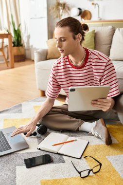 Genç, yakışıklı bir adam yerde rahatça oturur, fikirlerini çizerken dizüstü bilgisayarına odaklanır..