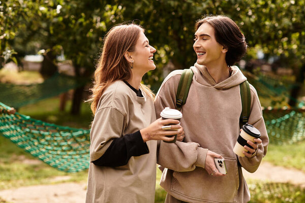 A young couple shares laughter while holding warm drinks, surrounded by green trees and nature.
