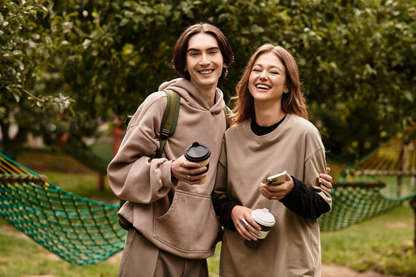 A young couple enjoys each others company in a beautiful outdoor space, smiling and holding coffee.