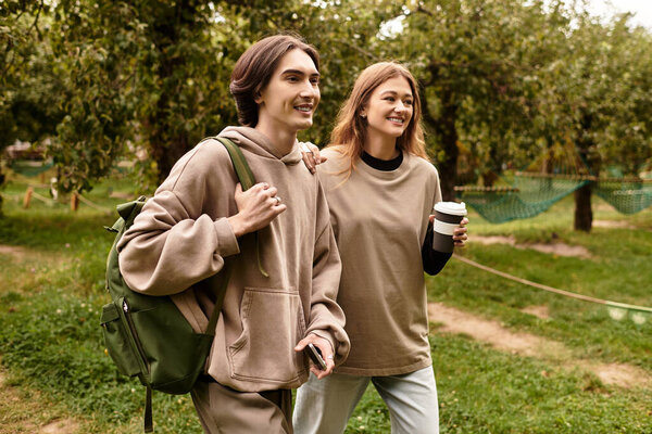 Young couple strolls through a scenic orchard, smiling and enjoying warm beverages together.
