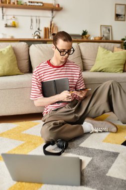 Genç adam yerde oturuyor, dizüstü bilgisayarının yanında çizim yapmaya odaklanmış..