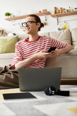 Genç bir adamın evinde rahat rahat otururken telefon ve dizüstü bilgisayar kullanması..