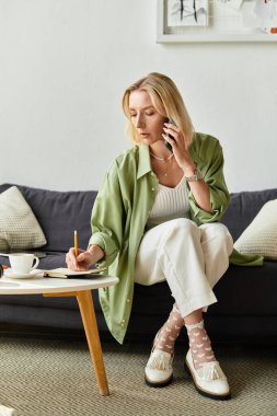 Günlük giysiler içinde bir kadın evde oturup, telefonla konuşuyor ve not alıyor..