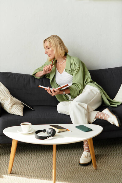 Beautiful woman in casual outfit sits on a couch, reflecting while holding a notebook and coffee.