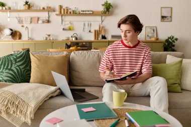 Genç bir adam evde ders çalışıyor, not alıyor ve şık bir ortamda dizüstü bilgisayarına odaklanıyor..