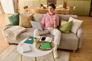 Odaklanmış genç bir adam, notlar ve dizüstü bilgisayarla çevrili şık dairesinden internetten ders çalışıyor..