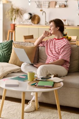 Genç bir adam rahat bir şekilde kanepede oturur, evde ders çalışmak için laptopunu kullanır, etrafı notlarla çevrilidir..