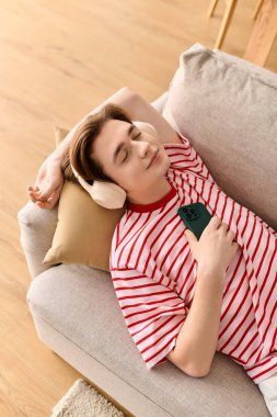 Genç bir adam kanepede uzanıyor, elinde telefonla internetten kulaklıkla ders çalışıyor..