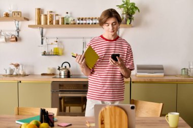 Genç bir adam canlı mutfağında telefon ve defter kullanarak online çalışmalarına odaklanıyor..