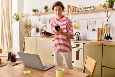 Genç bir adam şık bir mutfakta dizüstü bilgisayar ve telefon kullanarak ders çalışıyor..