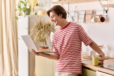 Genç bir adam internette dizüstü bilgisayarla çalışıyor. Evdeki mutfak tezgahına yaslanıyor..