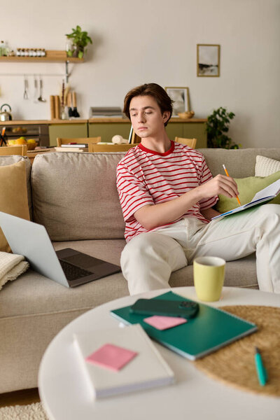 A young man studies online in his modern apartment, focused on his laptop with materials around him.