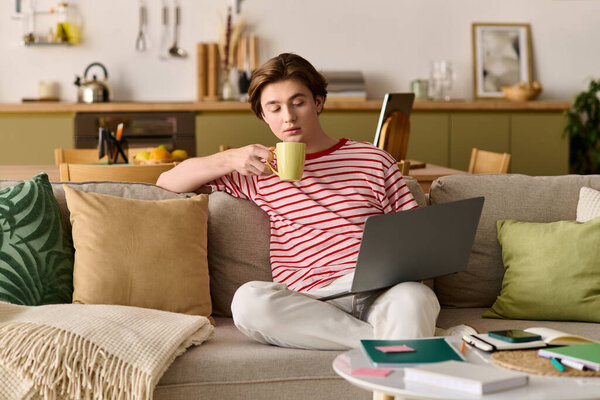 Молодой человек учится онлайн на удобном диване, наслаждаясь напитком в своей современной квартире.