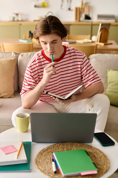 A young man engages in online studies at home, reflecting thoughtfully while taking notes.