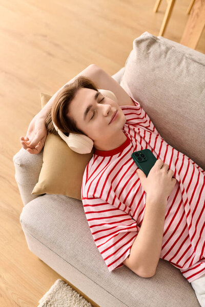 Молодой человек сидит на диване, учится онлайн с наушниками, телефон в руке.