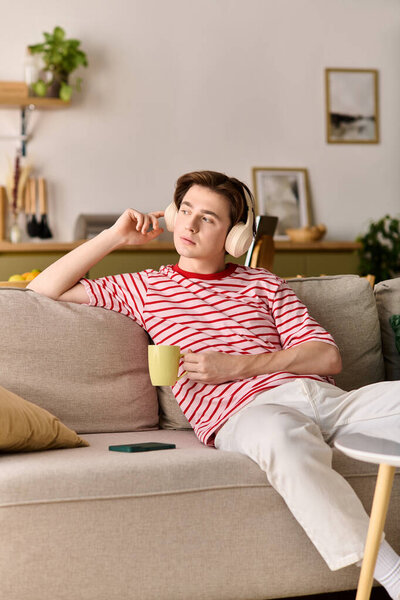 Молодой человек учится дома онлайн, расслабляется на диване в наушниках и выпивает.