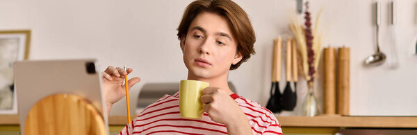 A young man is focused on his online studies while sipping coffee in his modern apartment, banner