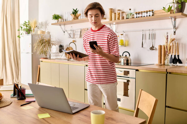 Молодой человек стоит на стильной кухне, учится онлайн с помощью ноутбука и телефона.